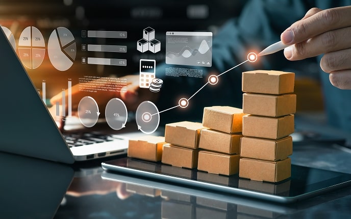 A hand points with a pen at a rising graph over stacked small boxes on a tablet, symbolizing business growth. Various digital charts and graphs are overlaid. A laptop sits beside the boxes on a glass desk, with blurred background lights.