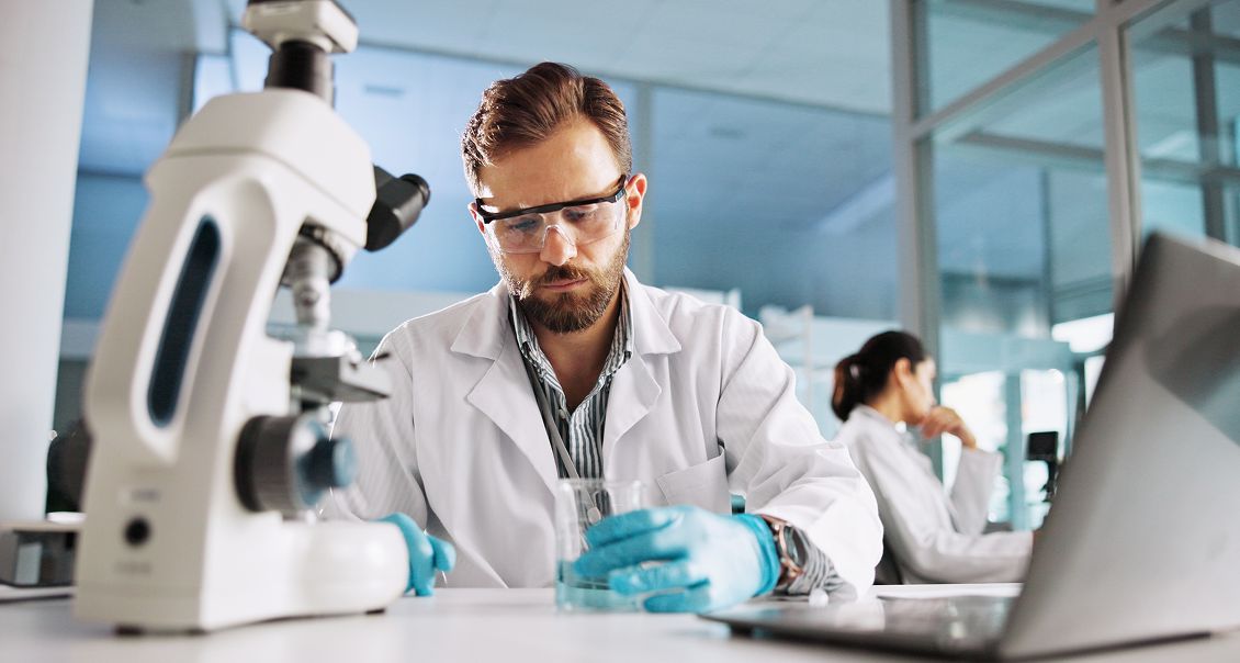 A male scientist wearing safety glasses and gloves examines a beaker at a lab desk beside a microscope. A laptop is open in front of him. In the background, another lab worker sits and talks on the phone. The lab is bright and modern with large windows.