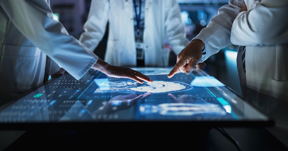 Three people in white lab coats examine and interact with a digital touchscreen table displaying detailed brain scans and medical data, suggesting a collaborative, high-tech medical or scientific setting. One person is pointing while another touches the screen.