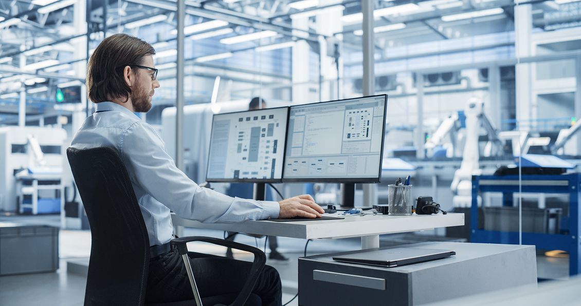 A man with long hair and glasses sits at a desk in a modern factory, working on two large computer monitors displaying technical diagrams and software. He is surrounded by industrial machinery and bright, spacious, high-tech surroundings.