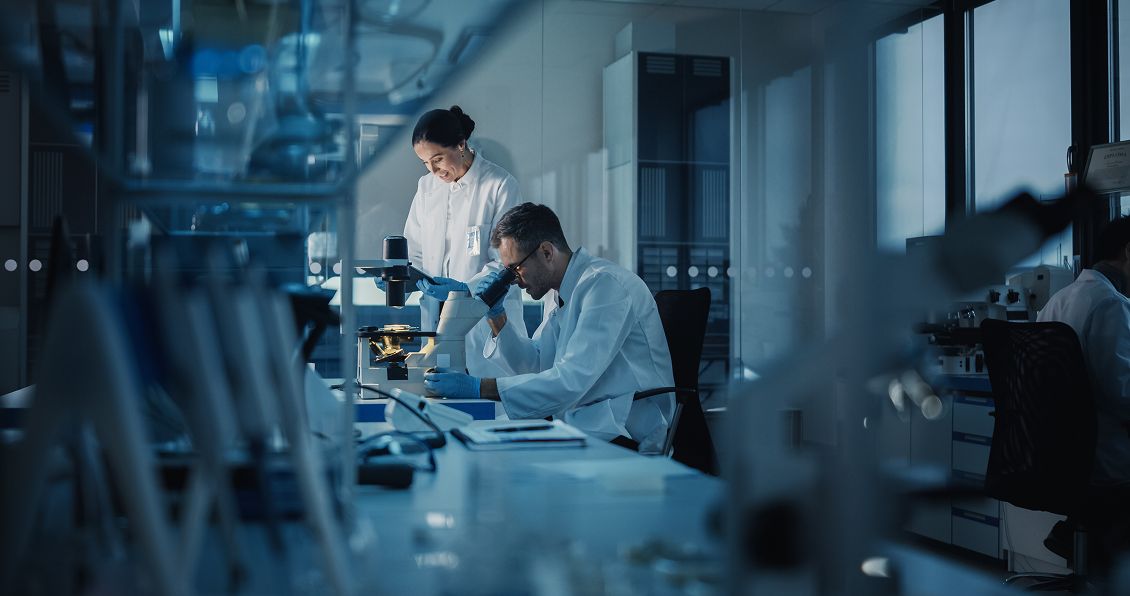 Two scientists in white lab coats work in a modern laboratory. One sits and examines a sample under a microscope, while the other stands nearby, smiling and observing. The lab is filled with equipment and bathed in cool blue-toned light.