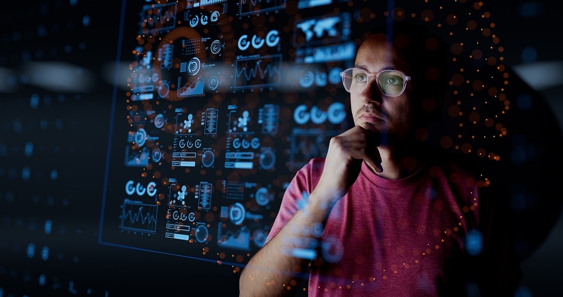 A man wearing glasses and a red shirt looks thoughtfully at a transparent digital screen displaying various charts, graphs, and data visualizations. The scene is dimly lit, emphasizing the glowing blue and orange graphics projected in front of him.