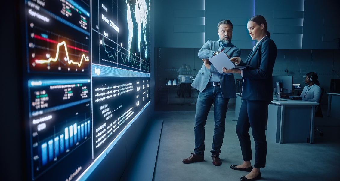 Two business professionals in formal attire discuss data on a clipboard while standing in a modern control room. A large wall screen displays graphs, world maps, and data analytics. Another person works at a computer in the background.