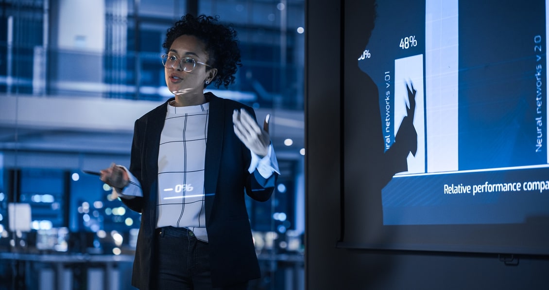 A person with short curly hair and glasses, wearing a black blazer and checked shirt, stands in a modern office and gestures while presenting a chart with performance statistics on a large screen. The chart shows bars with percentages, including one marked 48%.