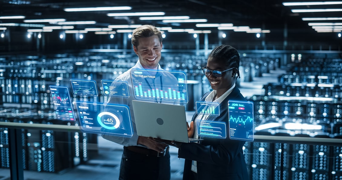 Two professionals stand in a modern data center, smiling at a laptop. Digital data graphics, including charts and analytics, appear as holograms above the laptop. Rows of servers and blue lights fill the background, creating a high-tech atmosphere.