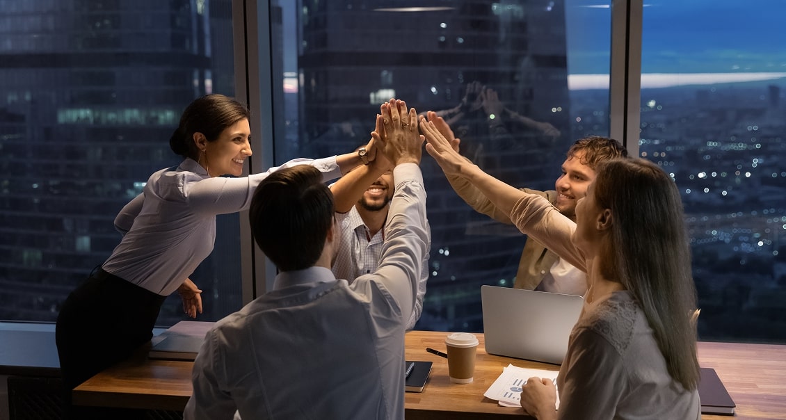 Four colleagues in business attire smile and high-five each other around a conference table in a modern office at dusk. Papers, a laptop, and a coffee cup are on the table. Large windows show a city skyline with buildings and lights in the background.