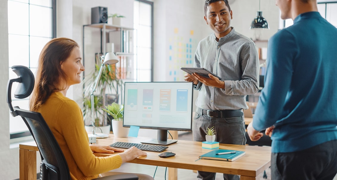 Three coworkers collaborate in a modern office. A woman sits at a desk with a computer displaying app designs, smiling at two standing men—one holding a tablet, the other gesturing. The office is bright with plants, shelves, and large windows.