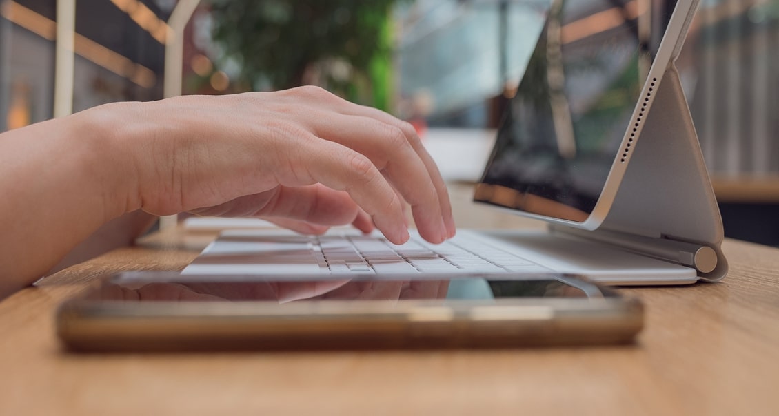 A close-up of a person’s hand typing on a white keyboard attached to a tablet with a foldable cover, placed on a wooden table. A smartphone lies face-up nearby. The background is softly blurred, suggesting a modern indoor workspace.
