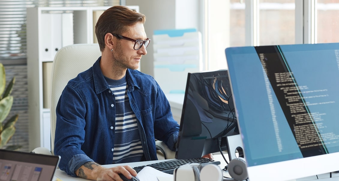 A man with glasses and a beard is sitting at a desk, working on a computer with code displayed on the monitor. He is wearing a blue jacket over a striped shirt. The office around him is bright and organized, with shelves and plants in the background.