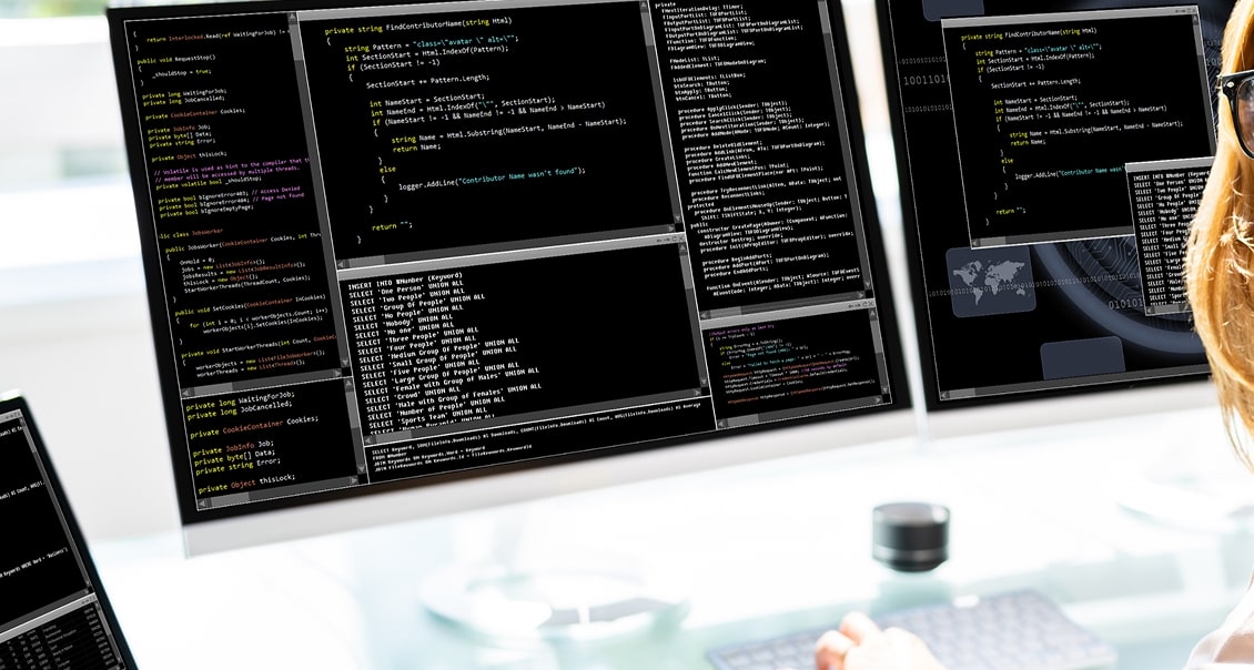 Two large computer monitors display multiple code editor windows with programming code and command-line interfaces. A person with long hair, wearing glasses, is seated in front of the screens, partially visible on the right side. A mouse sits on the white desk.