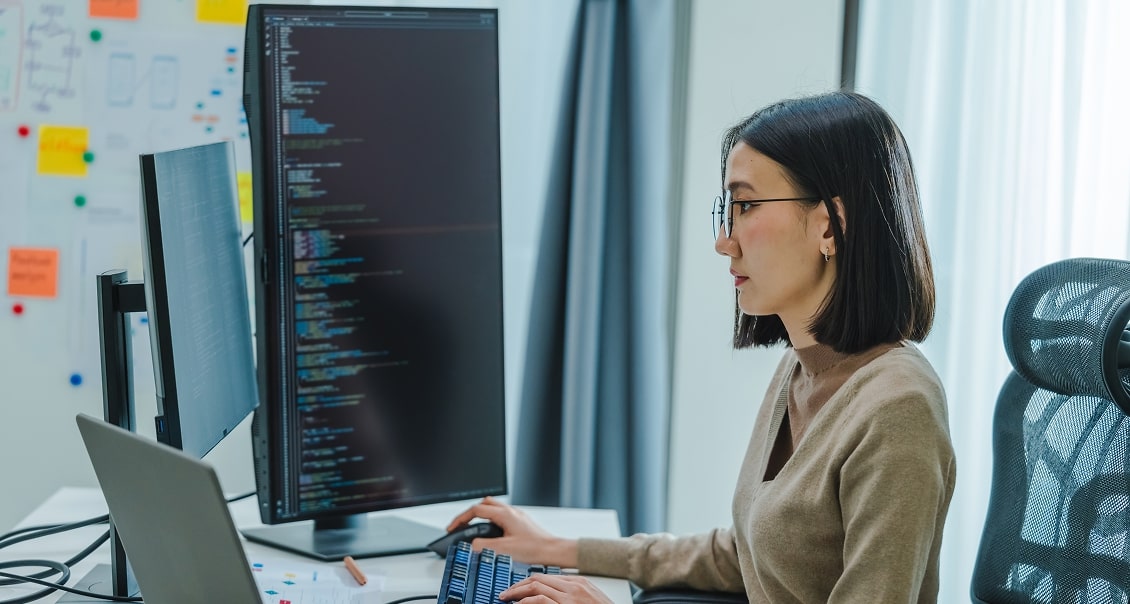 A woman with straight dark hair and glasses sits at a desk, coding on a large monitor. She uses a keyboard and mouse, with a laptop and second monitor beside her. Behind her are bright windows and a whiteboard with colorful sticky notes.