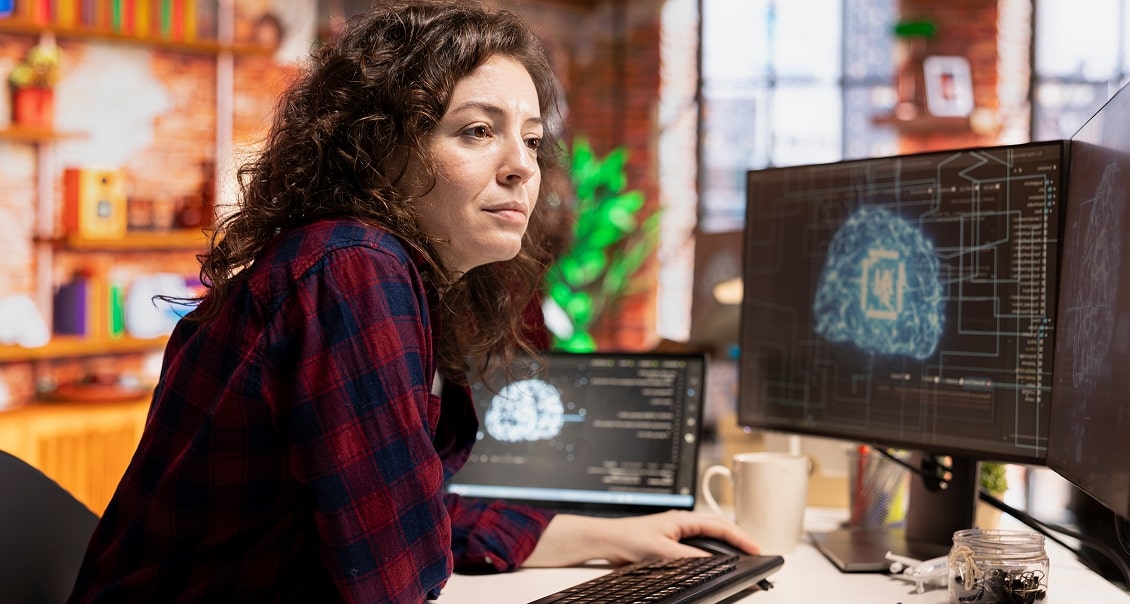 A woman with curly brown hair in a plaid shirt sits at a desk, looking at a monitor displaying a digital brain diagram and code. A laptop, coffee mug, and jar are on the desk. The background shows a cozy, colorful office with green plants.