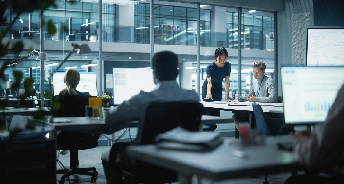 Modern office with glass walls at night; several people work at desks with computers. In the foreground, two colleagues collaborate at a desk, one sitting, one standing and pointing at papers. Other workers are focused on their screens in the background.