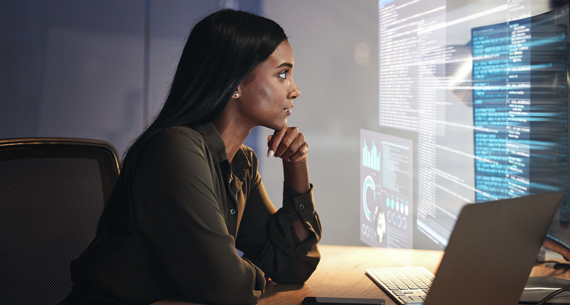 A woman with long dark hair sits at a desk, focused on a computer screen displaying code and data visualizations. She rests her chin on her hand, appearing thoughtful, with a laptop and paperwork on the wooden desk in front of her.