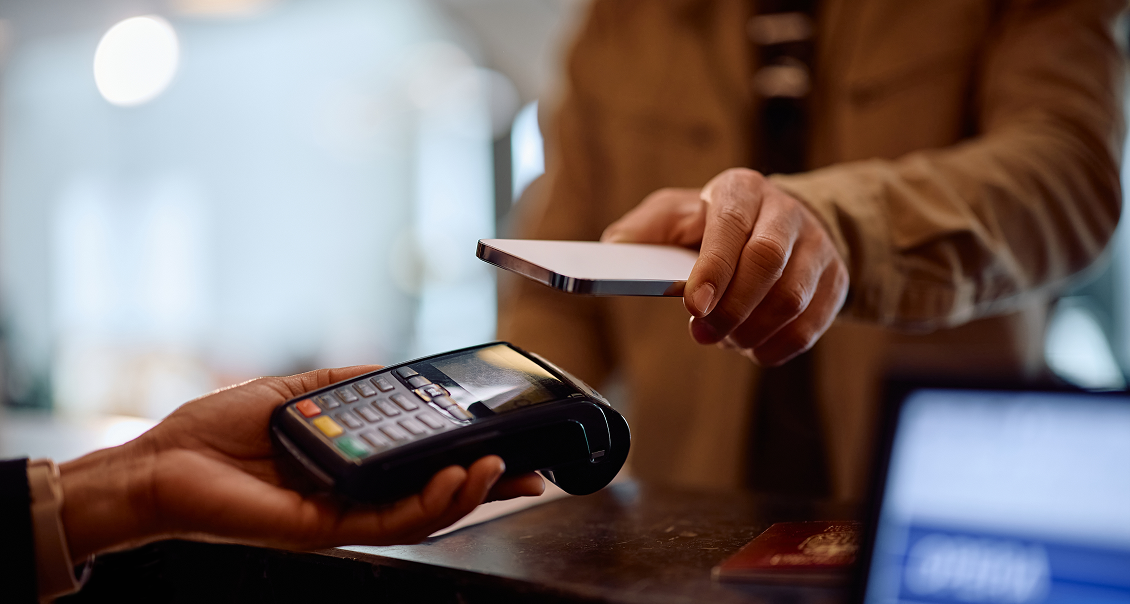 A person holds a smartphone over a card payment terminal for contactless payment. Another hand holds the point-of-sale device. The background is softly blurred, suggesting a retail or service counter setting.