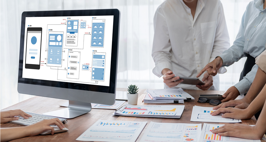 Four people collaborate at a desk covered with charts and documents. A computer screen displays wireframes for a mobile app. One person uses a tablet, while others point at papers and discuss designs, suggesting a UI/UX planning or development meeting.