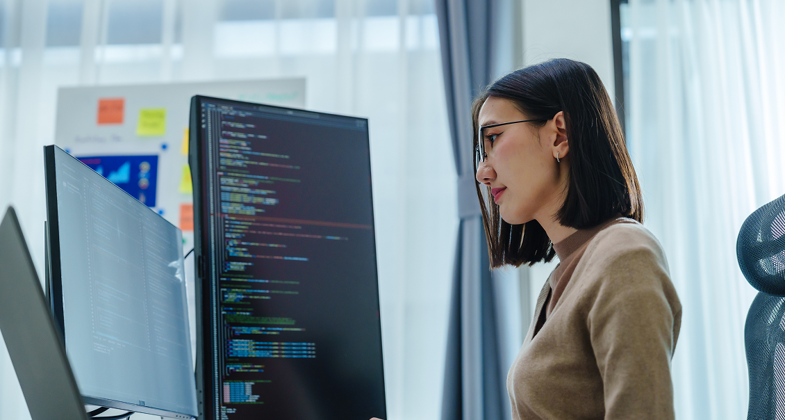 A woman with glasses and a brown sweater sits at a desk, focused on two large monitors displaying lines of colorful computer code. Behind her, there is a window with sheer curtains and a whiteboard with sticky notes in a modern office setting.