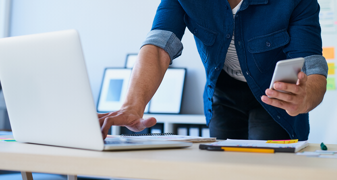 A person stands at a desk, holding a smartphone in one hand and using a laptop with the other. Notebooks, papers, and pens are spread on the desk. The person wears a blue shirt over a striped t-shirt. The background is an office setting.