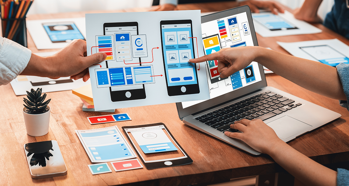 A team collaborates around a desk with laptops, smartphones, and printed UI mockups. One person holds up a paper showing mobile app wireframes, while another points at the designs, discussing app development and user interface layout.
