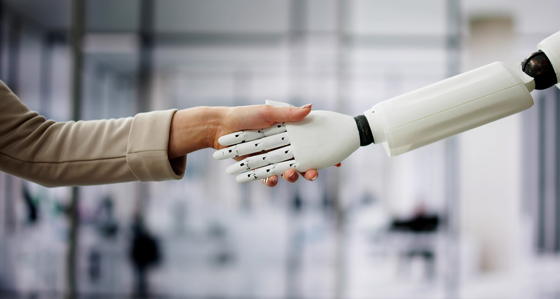 A human hand in a beige sleeve shakes hands with a white robotic hand. The background is blurred, suggesting a modern office or business setting, symbolizing collaboration between humans and artificial intelligence or technology.