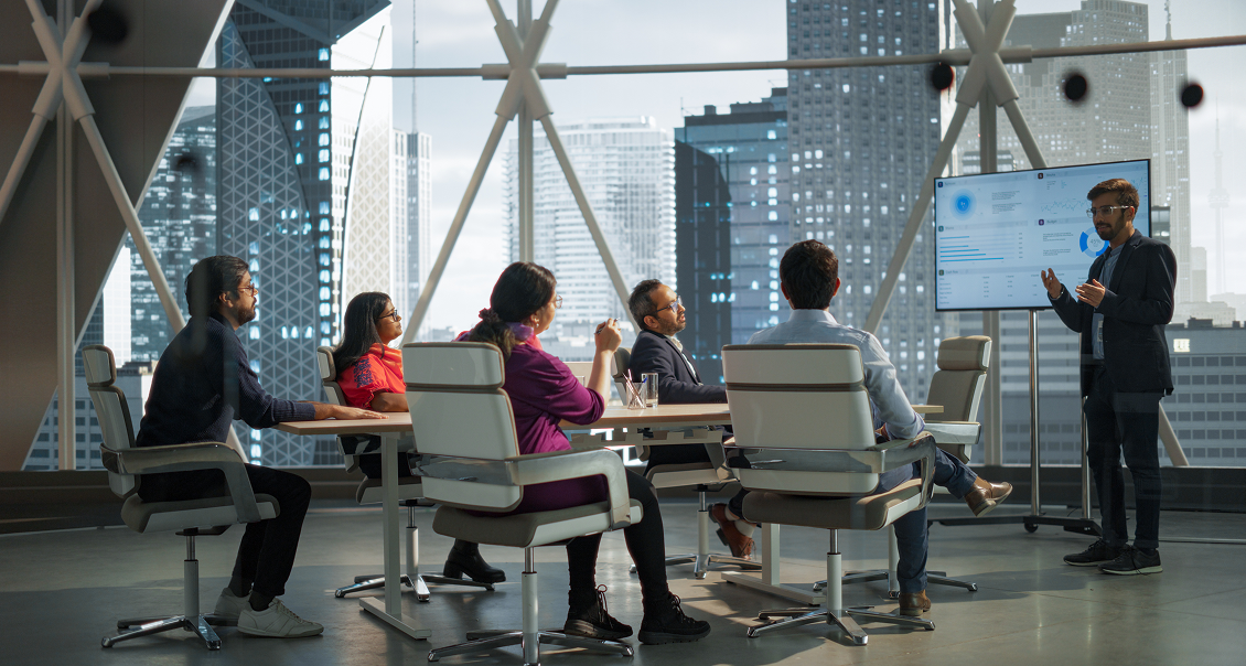 Six people sit around a conference table in a modern office with large windows showing a city skyline. One person stands by a screen, presenting data to the group, who are listening attentively. The atmosphere appears professional and collaborative.