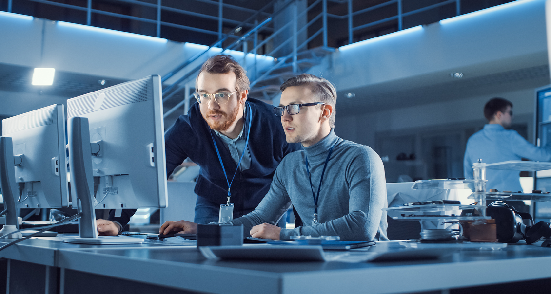 Two men wearing glasses and security badges work together at a desk with dual computer monitors in a modern, high-tech office with blue lighting. One man sits typing while the other stands, leaning in to look closely at the screen. Other workers are in the background.