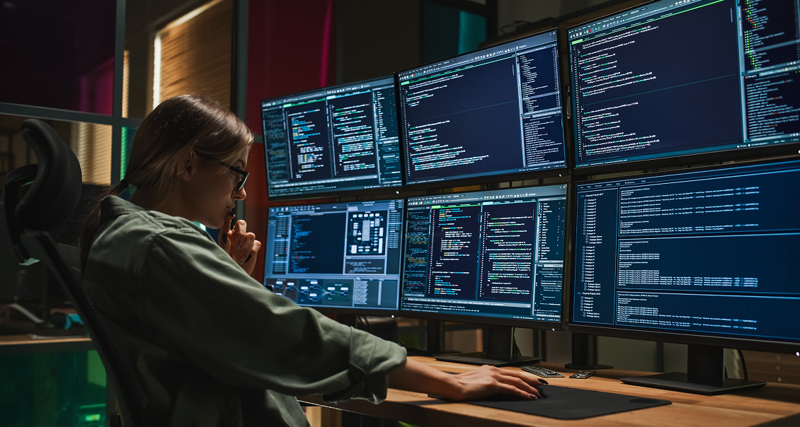 A person sits at a desk in a dimly lit room, focused on five large computer monitors displaying lines of colorful code and data. They rest their chin on one hand, wearing glasses and a green shirt, suggesting intense concentration on programming work.