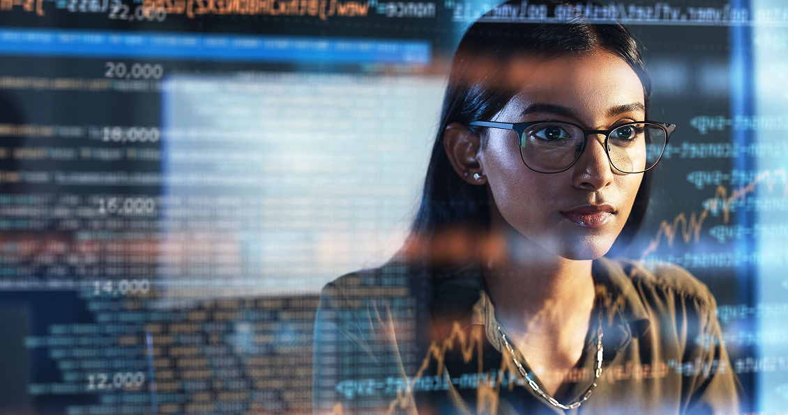 A woman with long dark hair and glasses looks intently at a computer screen displaying colorful code and financial graphs, reflecting on her face. She is focused, sitting in an office with digital data overlays in the foreground.