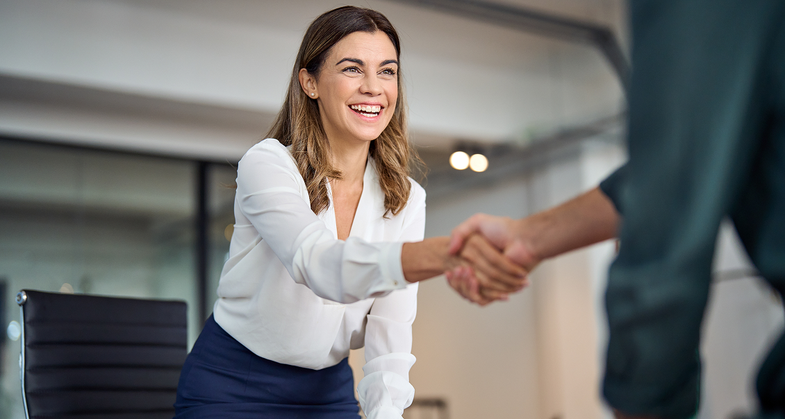 A smiling woman with brown hair, wearing a white blouse and blue skirt, stands and extends her hand to shake hands with another person. They are indoors in a modern office setting with glass walls in the background.