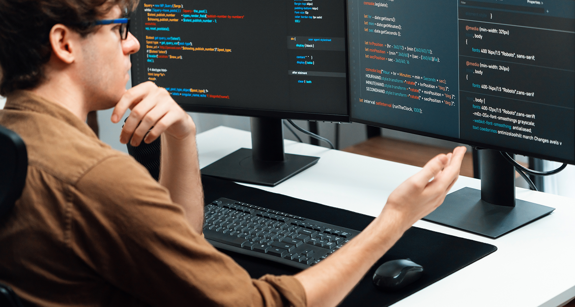 A person wearing glasses sits at a desk with a keyboard and mouse, facing two large monitors displaying colorful programming code. The person appears focused, with one hand on their face and the other gesturing toward the screens.