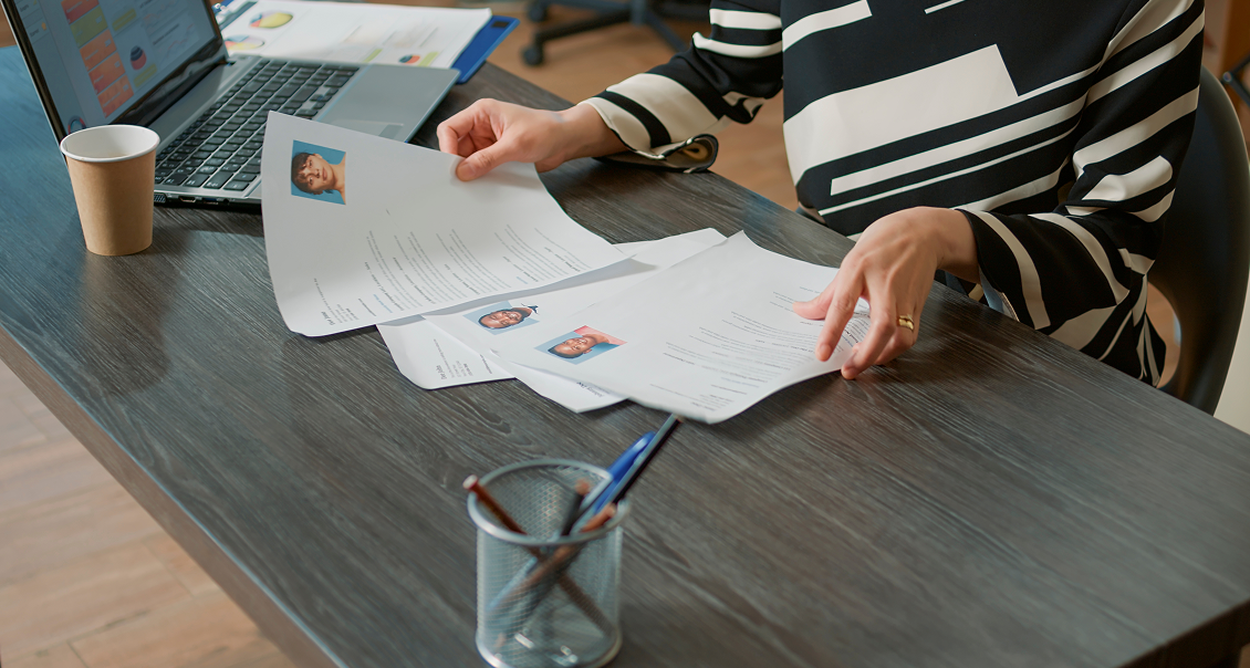 A person in a black-and-white striped top reviews printed resumes with headshots at a wooden desk. A laptop, a paper coffee cup, and a pen holder with pens are also on the desk. The scene suggests a hiring or interview process.