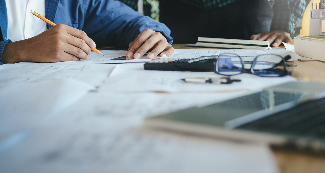 Two people work at a desk covered with architectural blueprints, a calculator, eyeglasses, and a laptop. One person writes with a pencil on a document, while another stands nearby holding a notebook. The scene suggests collaboration and planning.
