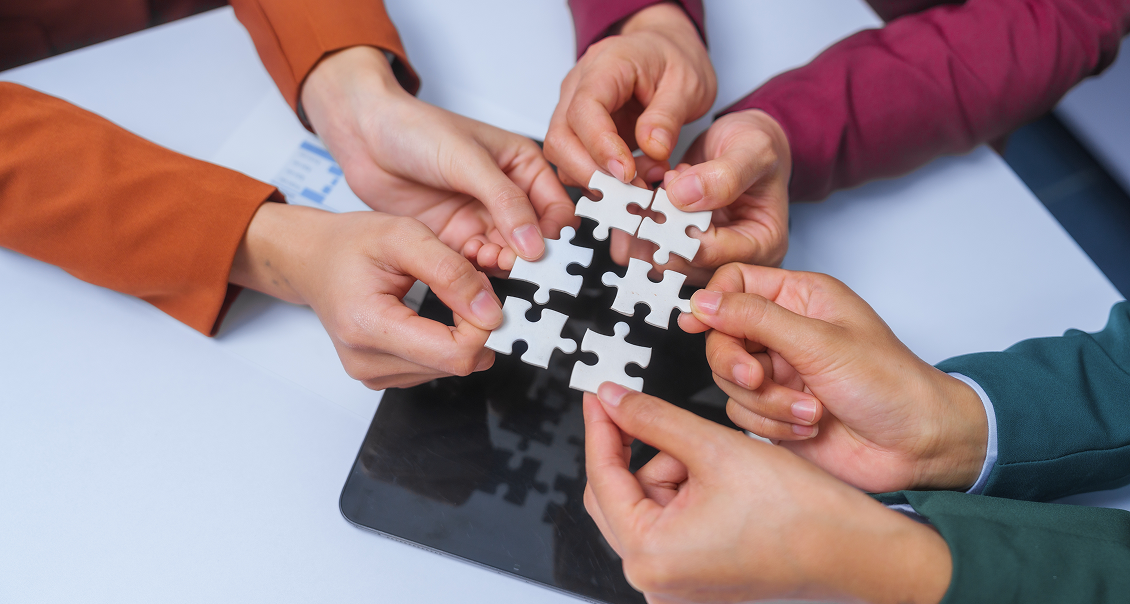 Six hands, each wearing differently colored sleeves, work together to connect four white puzzle pieces over a black tablet on a white table, symbolizing teamwork and collaboration.