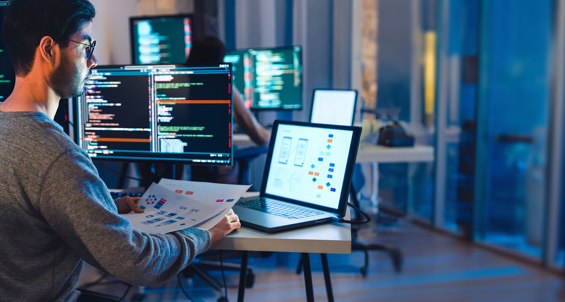 A man sits at a desk with a laptop displaying app wireframes and holds printed diagrams. Multiple monitors display code. The office has large windows, a blurred cityscape outside, and another person working in the background. The lighting is cool and dim.