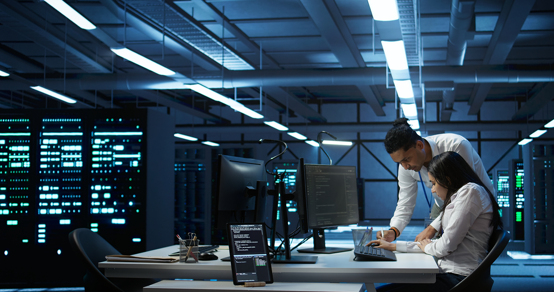 Two people in business attire work together at a desk with multiple monitors and a tablet displaying code in a modern, dimly lit server room filled with tall racks of servers and blue lighting. One person is seated typing while the other points at the screen.