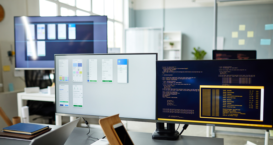 Two computer monitors on a desk in a modern office; one displays app interface designs, the other shows code in a dark terminal. In the background are a large screen, glass walls, sticky notes, and sunlight streaming through windows.