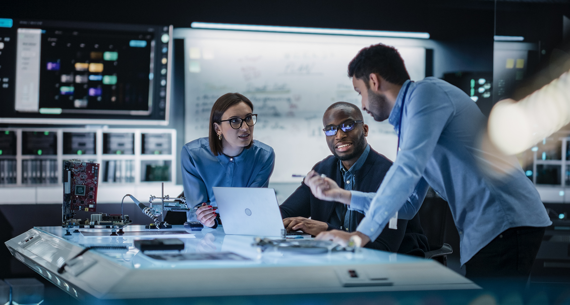 Three engineers sit together at a high-tech workstation with a laptop, circuit boards, and monitors. Two listen attentively while a third, standing, gestures as he talks. Large screens and technical equipment fill the modern lab’s background.
