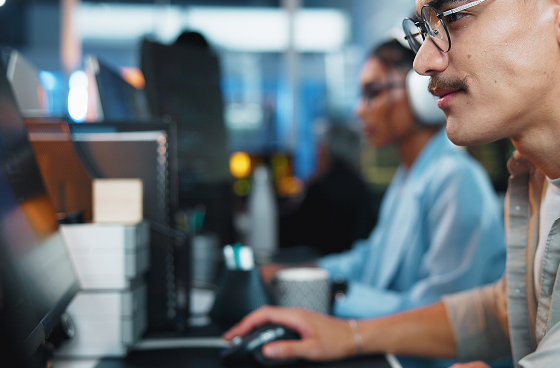 A man wearing glasses works at a computer, using a mouse, with a mug nearby. In the background, a woman with headphones and glasses also focuses on her computer. The office setting appears modern and slightly blurred in the background.