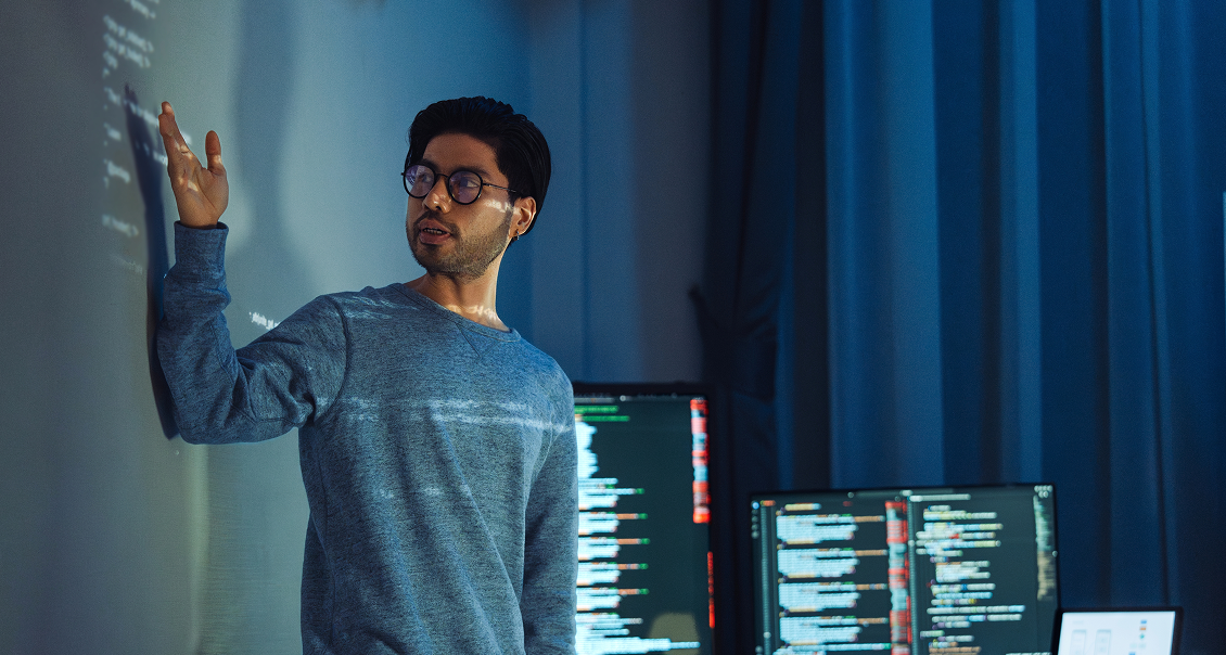 A man wearing glasses and a gray sweater points at code projected on a wall. Behind him are monitors displaying more code. The room is dimly lit with blue tones, suggesting a tech or programming environment, possibly during a presentation or meeting.