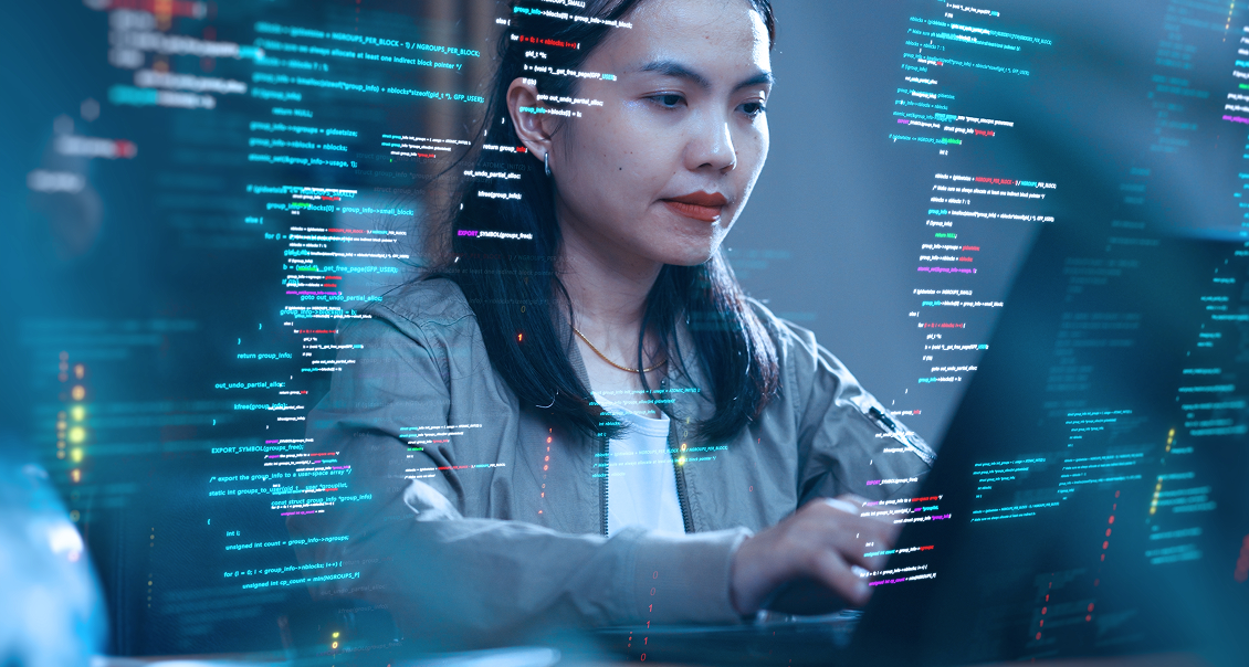 A woman with long dark hair types on a laptop, focused. Colorful lines of programming code are digitally overlaid around her, suggesting she is coding or working in software development. The background is blurred, emphasizing her concentration.