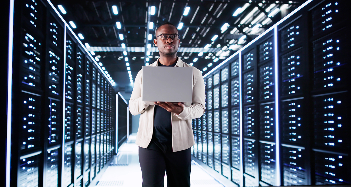 A person wearing glasses and a light jacket stands in a brightly lit server room, holding an open laptop. Rows of server racks with blinking lights line both sides, and the modern room has futuristic ceiling lights.