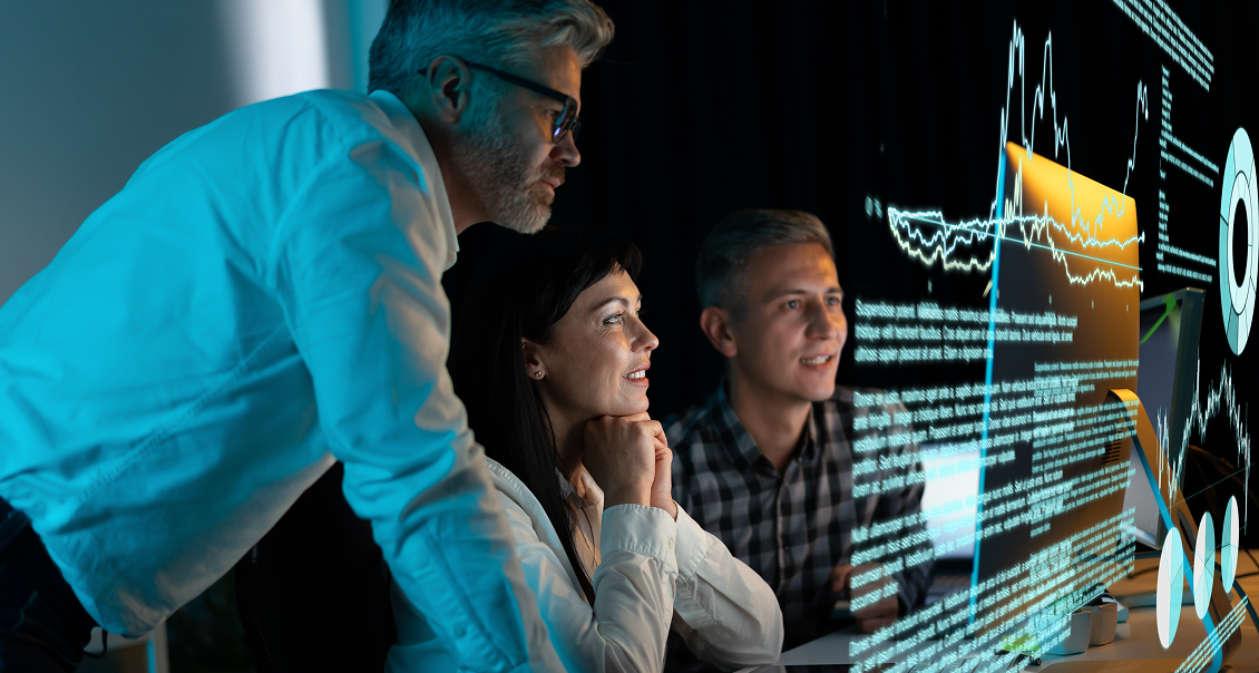 Three professionals, two men and one woman, sit and stand in a dark office, focused on a large computer screen displaying glowing data charts, graphs, and code. The group appears engaged and collaborative, working together on a high-tech project.