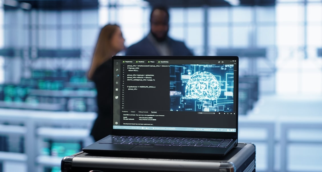 A laptop displaying code and a digital brain graphic sits on a cart in a modern, high-tech office. The blurred background shows two people talking and large windows letting in natural light, emphasizing a professional, tech-focused environment.