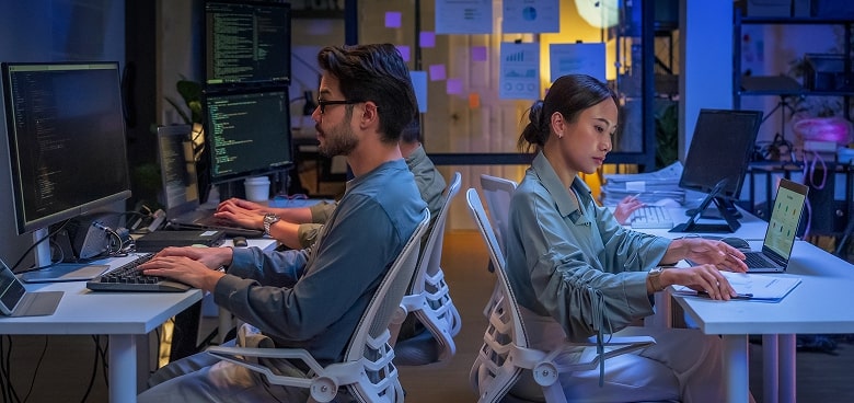 Two people sit back-to-back at desks, focused on their work. The man types on a keyboard with code on his screen, while the woman writes notes, referencing her laptop. The office is modern, with multiple monitors and blue-toned lighting.