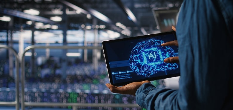 A person holds a tablet displaying a glowing blue digital brain labeled AI while standing in a modern, high-tech server room filled with racks of computer equipment and blurred lights in the background.