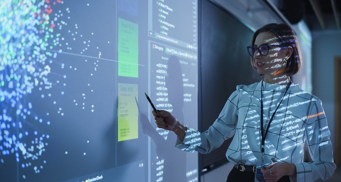 A woman in glasses stands in front of a large digital screen displaying colorful data visualizations and code. She points to the screen with a pen and smiles, wearing a white blouse in a modern office or classroom setting. Light from the screen reflects on her.