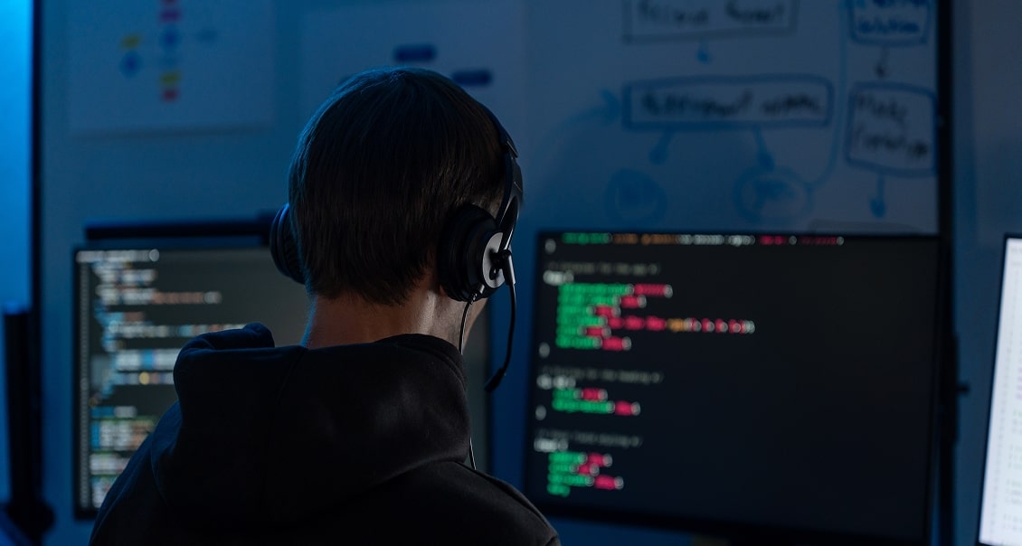 A person wearing headphones sits at a desk with multiple monitors displaying lines of code. The room is dimly lit, with blue light illuminating the scene. In the background, a whiteboard with diagrams and notes is visible. The person is seen from behind.