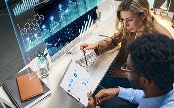 Two professionals sit at a desk analyzing data. A woman looks at graphs on a large screen, while a man points at a tablet displaying charts. Both appear focused, surrounded by office items like notebooks and a lamp. Blue digital data visualizations overlay the image.