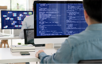 A person sits at a desk in front of a large computer monitor displaying lines of code. Another person works at a computer in the background, where a flowchart or network diagram is visible. Books and a smartphone are on the first desk.