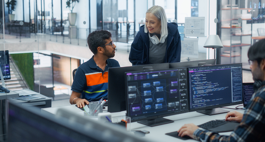 Three people work in a modern office with large windows. Two—one man in glasses and a striped polo, and a woman with blond hair and headphones—are talking. Computer monitors display code and diagrams. Another person types at a nearby desk.
